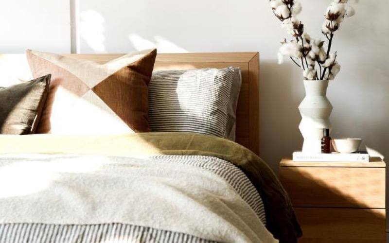 swath of sunlight shines across a well made bed and bedside table with a vase of cotton plants